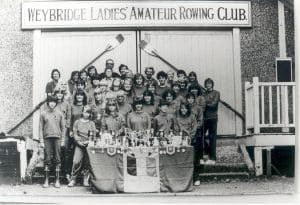 A group of women stand in front of Weybridge Ladies Amateur Rowing Club clubhouse. A table with trophies is in front of them.