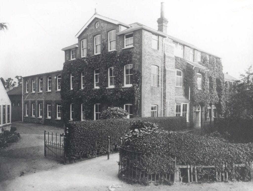 Black and white photograph of exterior view of the Claudius Ash Sons & Co. Ltd. works in Churchfield Road, Walton, showing ivy clad building behind neatly clipped hedge, c.1914.