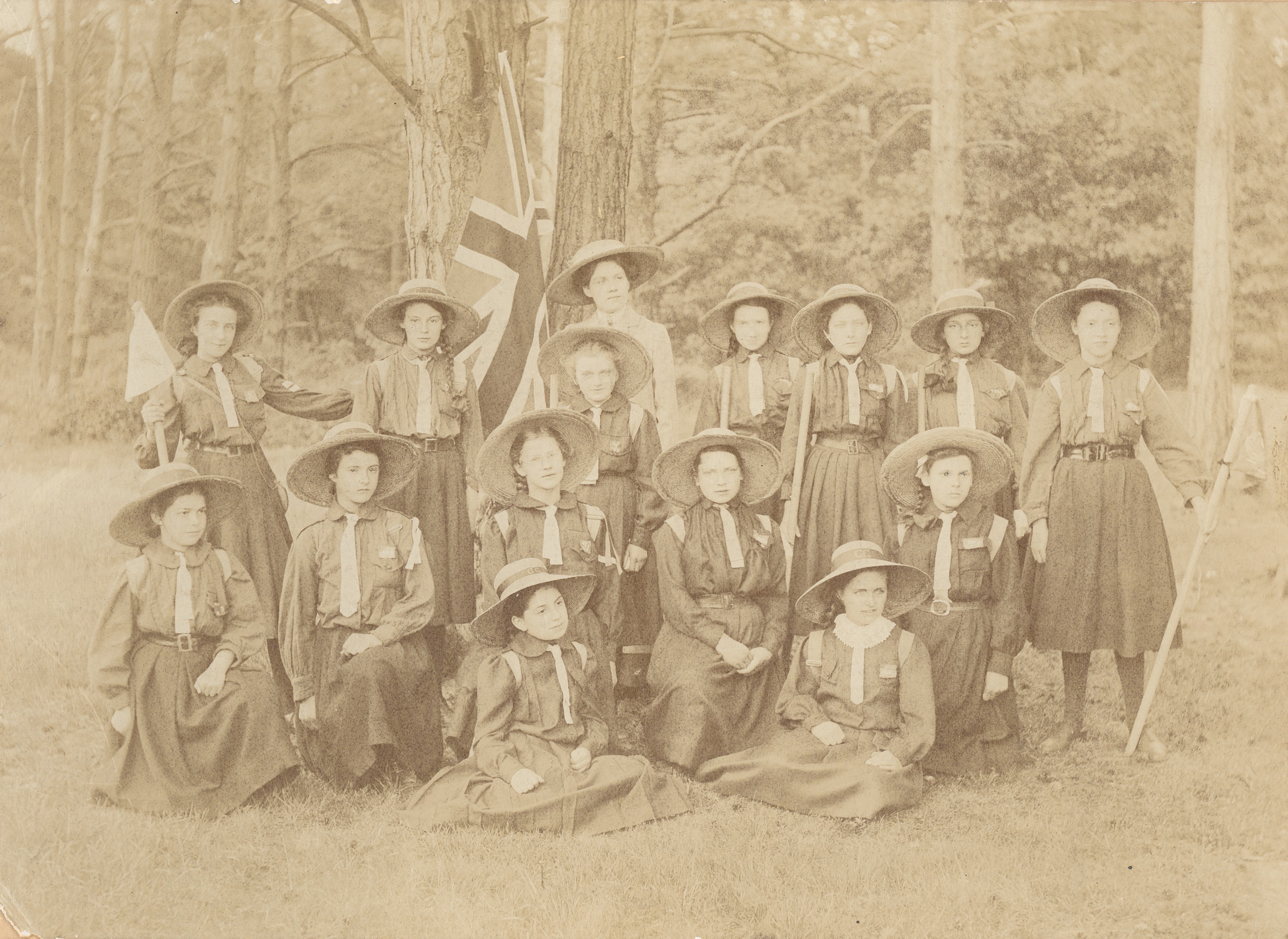 The 1st Weybridge Girl Guides, 1910, at Brooklands Estate. The group of girl guides are seated and standing in possibly the grounds of Brooklands House. The Union Jack is held by Miss James (Captain).