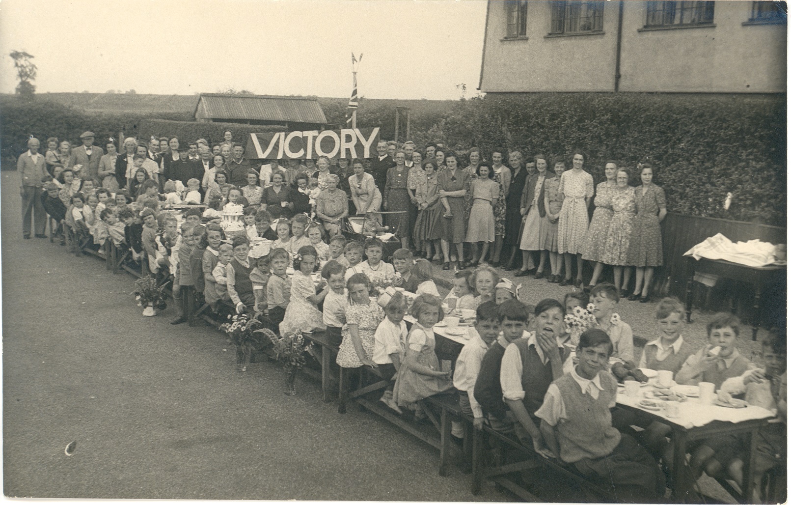 Photograph of Victory celebrations probably in Florence Road, showing a long table with children sitting at it and adults in the background holding a 'VICTORY' banner.