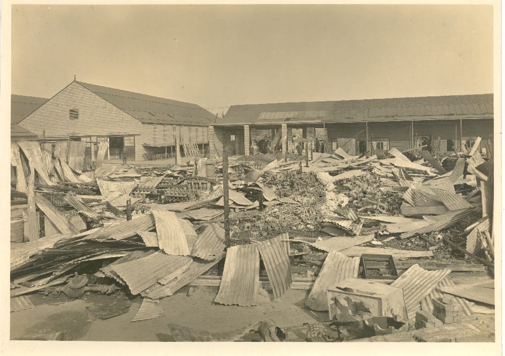 Image of factory showing sheets of corrugated iron strewn around and a few upright very charred posts.