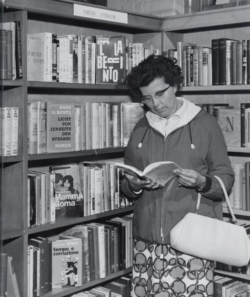 Interior of Walton Library, showing a member of the public looking at a book in a foreign language in front of a bookcase labelled 
