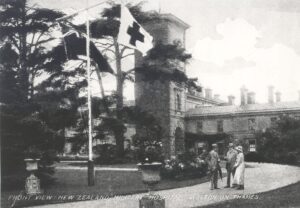 A view of Mount Felix mansion when it was being used as a military hospital for New Zealand soldiers from 1915-19. The view is from the driveway, flanked by large flower-vases, looking towards the building. A flag staff bears the flags of New Zealand and the Red Cross, and a group of three people stand in the foreground.