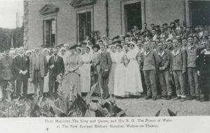A postcard published to commemorate the visit of King George V, Queen Mary and Edward, Prince of Wales, to the New Zealand Military Hospital at Mount Felix in 1917. The King is stood fourth from the left in the front row, with the Queen stood to his right. Many of the soldiers and nurses at the hospital are stood around them.