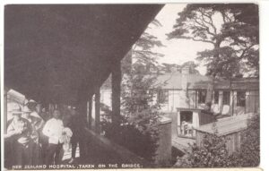 This postcard of Mount Felix is entitled 'New Zealand Hospital, taken on the bridge.' A ward orderly and several troops stand in the left foreground on the covered bridge, from which there is a general view of the hospital buildings on the right, including the 'Lord Plunket Shelter'.