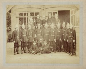 222.1969 Photograph of Weybridge Fire Brigade grouped on and around the 1879 Fire Engine outside main doors of fire station in 1880s or early 90s. Five Men on engine. Ten men standing in front, two boys sitting on the ground, small table with trophies on it in center of group