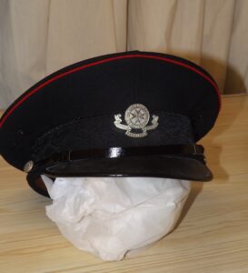 St. John's Amulance hat, black worsted with red piping round the top of the brim. Black silk braiding of oak leaves and acorns around the hat, with silver metal St. John's Ambulance Brigade badge on front. Patent leather narrow strap over peak with silver St. John's star buttons either side. Black shiny peak, lined underside with green plastic. Inside hat lined with leather, stamped "Real Brown Leather". Mica covering to crown.