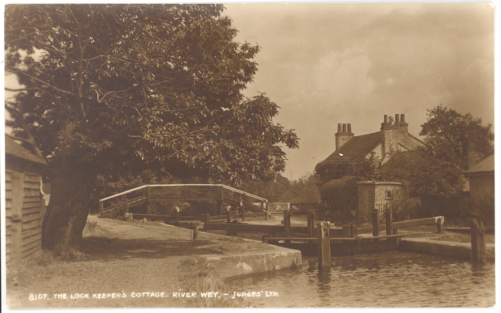Copy of Sepia postcard of The Lock Keeper's Cottage, River Wey, Weybridge, c. 1930s with a footbridge in the background. The lock keeper's cottage is on the right and to the left the edge of the stable for the horses that towed the barges is just visible next to a very large tree in full leaf.